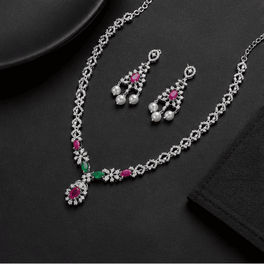 Luxury lifestyle display of the Vrindae Argente sterling silver ruby and emerald CZ necklace set with matching pearl earrings on a grey and silver premium background.