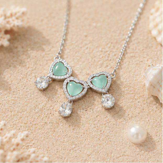  Beach-inspired flat lay of the Vrindae Argente Ocea Heart sterling silver necklace and earrings with mint green heart stones.