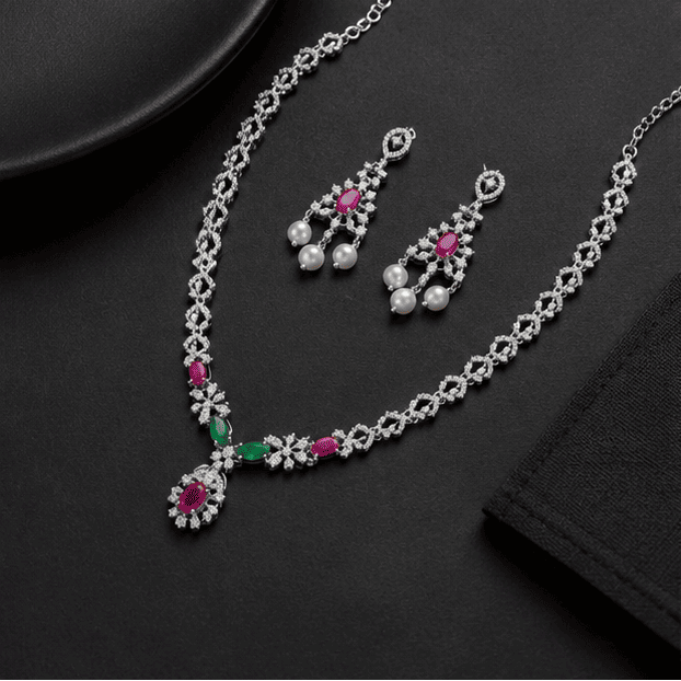 Luxury lifestyle display of the Vrindae Argente sterling silver ruby and emerald CZ necklace set with matching pearl earrings on a grey and silver premium background.